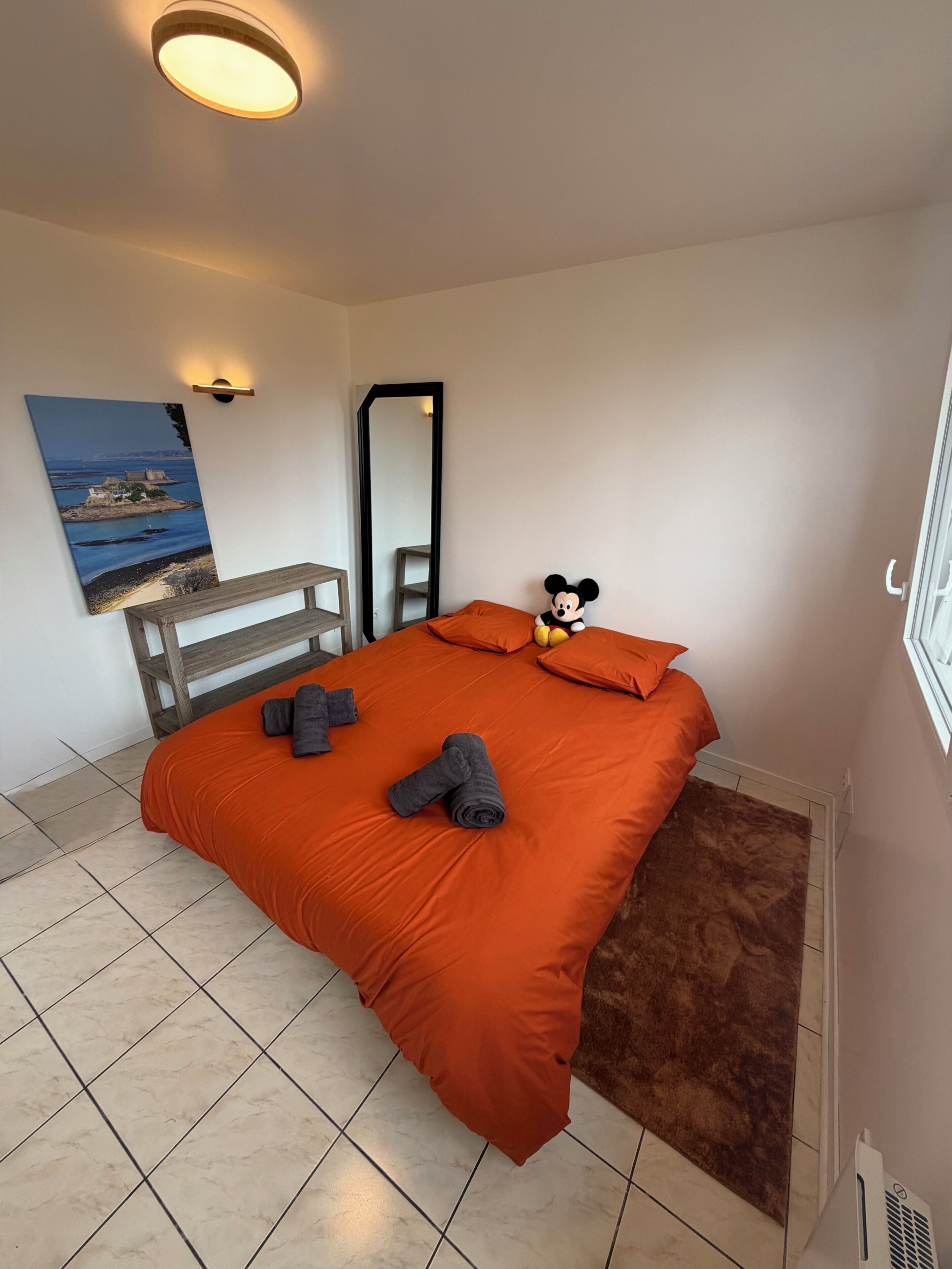Une chambre à coucher avec un lit, un doudou à l’effigie de Mickey Mouse, un tableau, un miroir et un banc en bois.