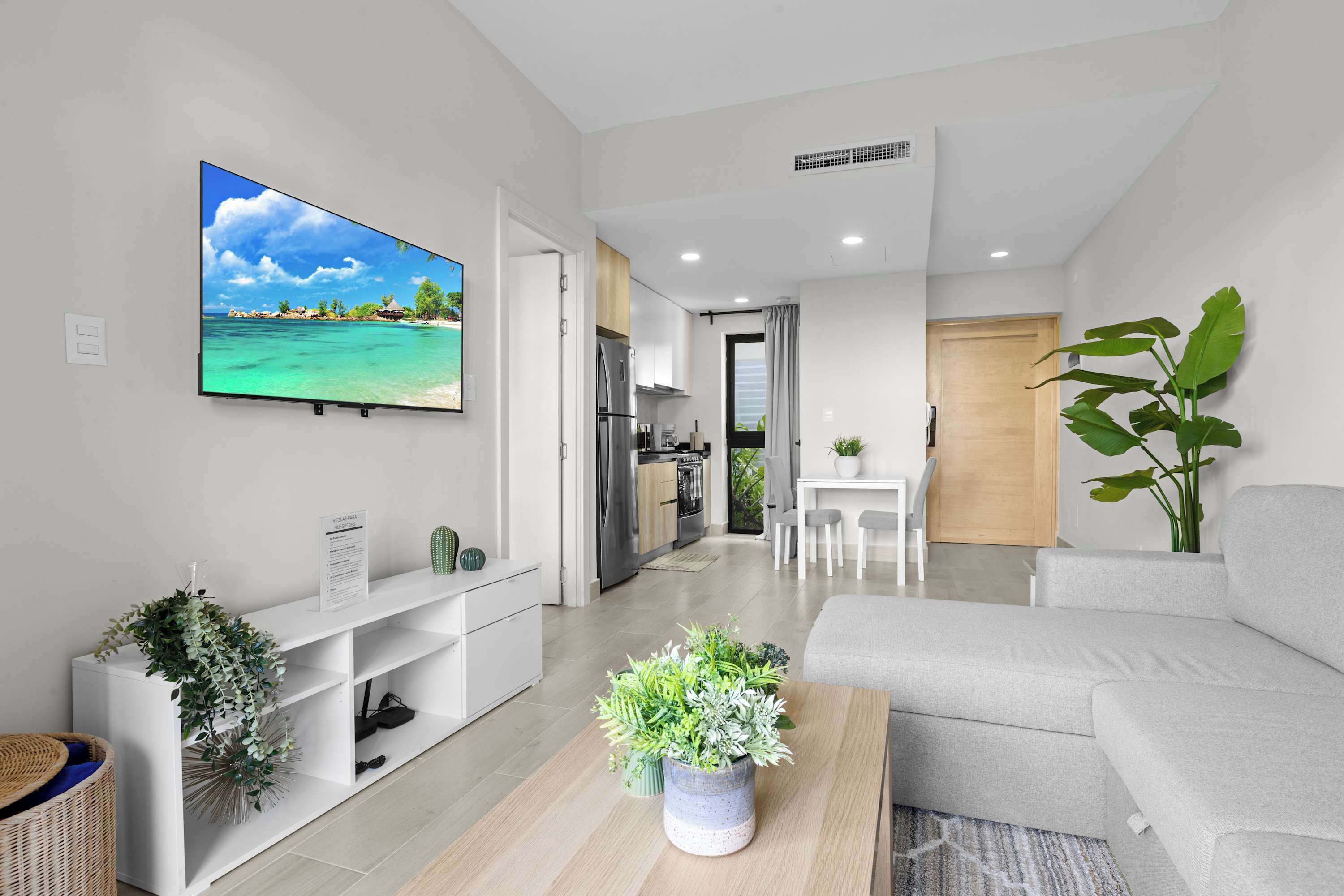 A modern living room with a flat-screen TV, a sofa, a coffee table with plants, and a kitchen area in the background.