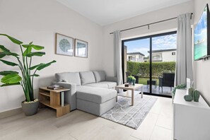 Ein modernes Wohnzimmer mit einem grauen Sofa, einem Couchtisch, einer großen Topfpflanze und einer Glastür, die auf einen Balkon führt.
