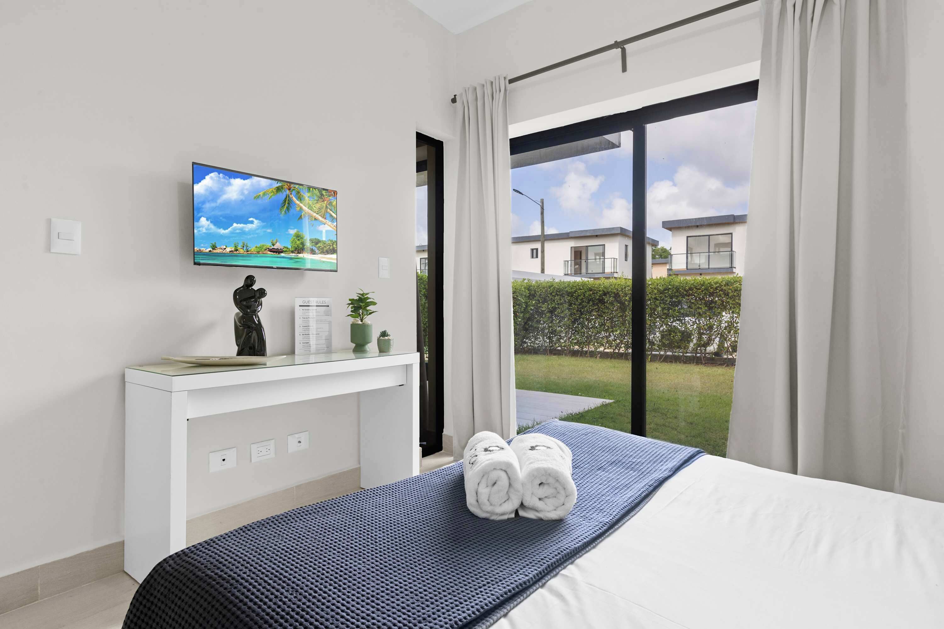 Hotelkamer met een bed, een flatscreen televisie aan de muur, een klein bureau met een plant en uitzicht op een woongebied door een glazen deur.