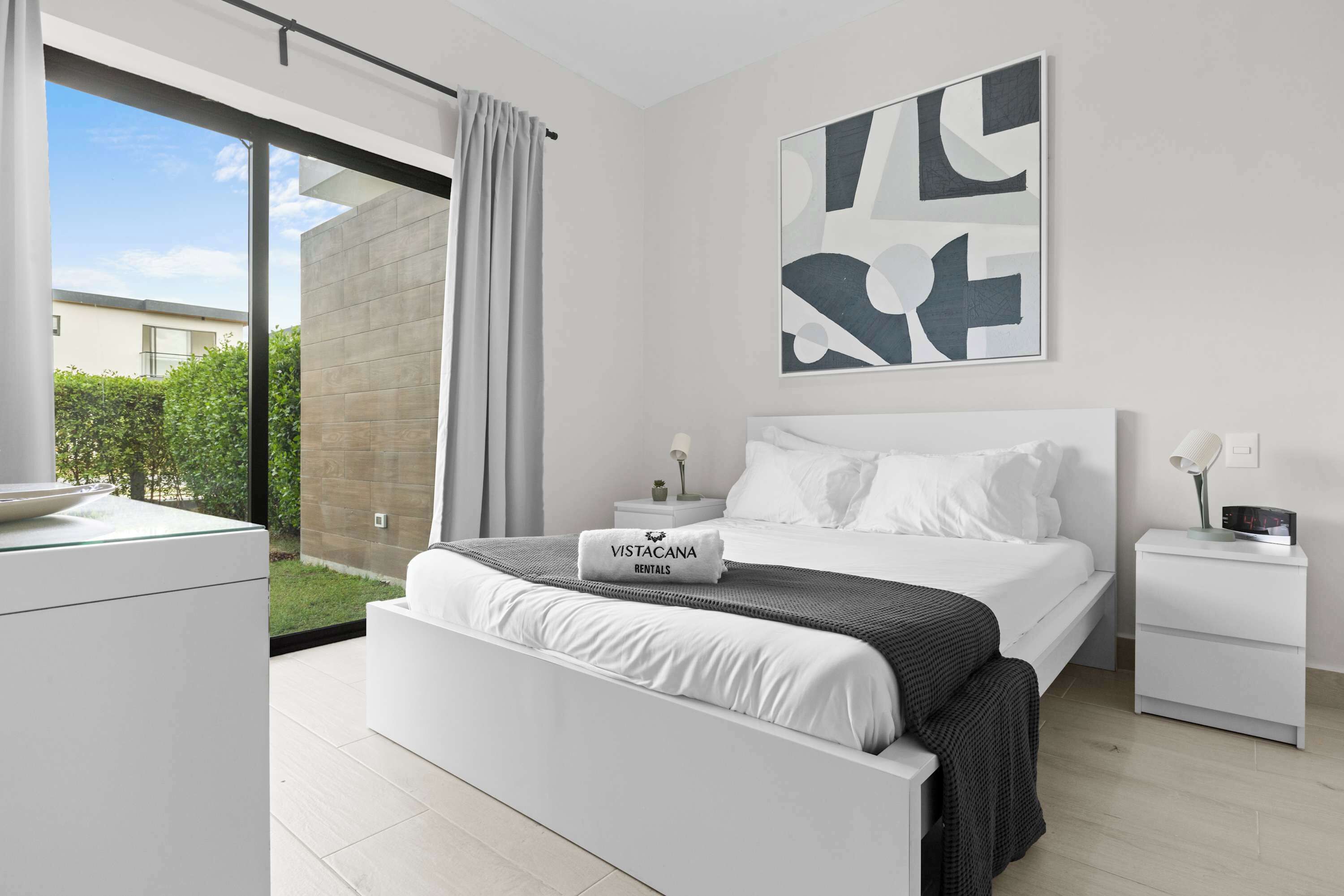 A modern bedroom with a large bed, a nightstand, a digital clock, and a view of a garden through a sliding door.