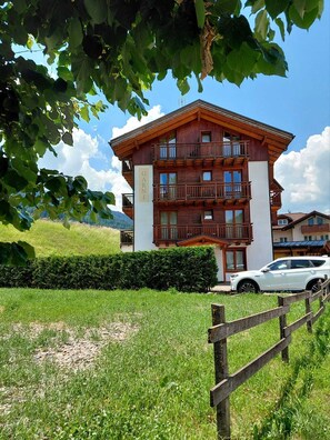 A multi-story wooden building with balconies, a white car parked in front, and a hedge in the foreground. - ABETE (Mestriago)