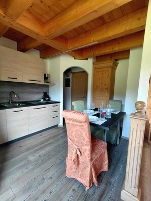 A kitchen with wooden cabinets, a dining table set for two, and a chair with a patterned cover. - ABETE (Mestriago)
