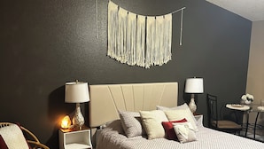 A bedroom with a bed, bedside tables, a chair, a rug, and a wall hanging. - Private Spa Suite + Patio
The Tranquil Corner (Tucson)