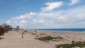 Uma praia com céu claro, terreno arenoso e edifícios ao fundo.