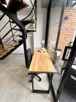 Ein moderner Arbeitsbereich mit einem Holztisch, einer schwarzen Metalltreppe und Blick auf ein Ziegelgebäude durch eine Glastür.