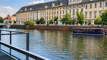 A riverside view of a historic building with a prominent tower, a boat docked on the river, and a modern bridge railing in the foreground.