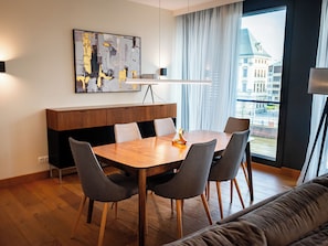 A modern dining area with a wooden table and grey chairs, a painting on the wall, and a view of buildings through a window.