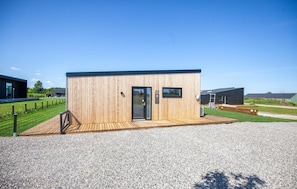 Ein kleines Holzgebäude mit Tür und Fenstern, umgeben von Kies und Gras.