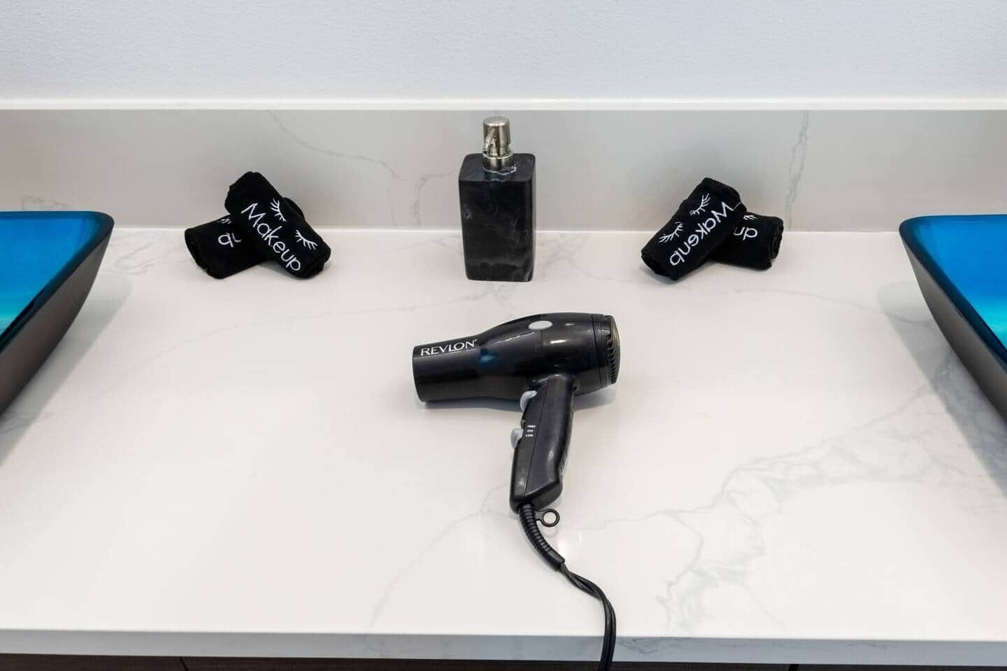 Ein Badezimmer mit weißer Ablage, einem schwarzen Föhn und zwei schwarzen Flaschen mit weißer Beschriftung.