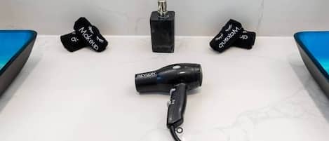 Casa de banho com bancada branca, secador de cabelo preto e duas embalagens pretas com texto branco.