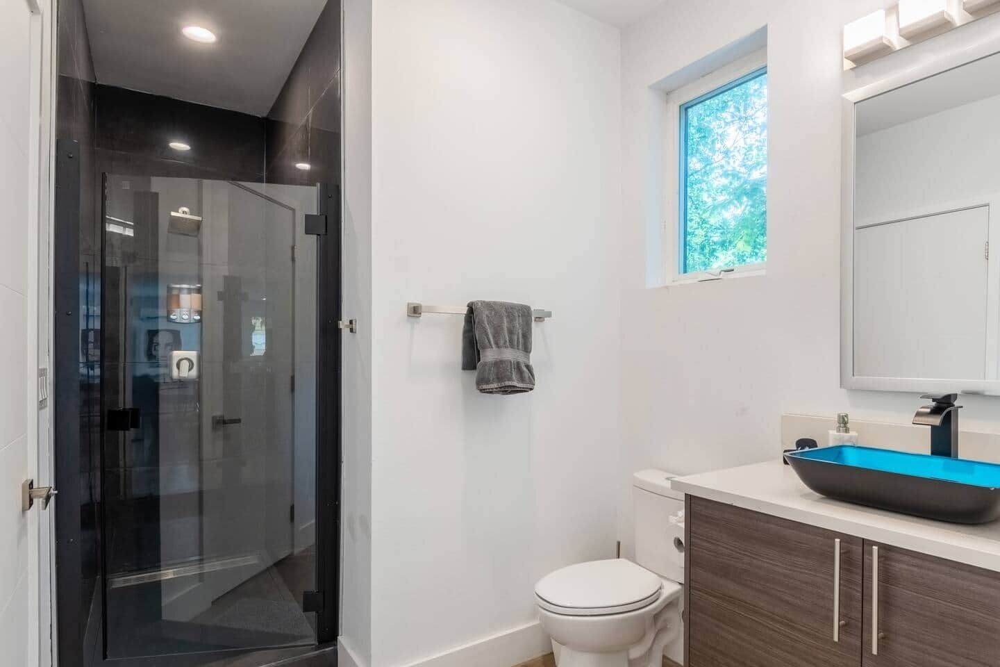 Une salle de bain moderne avec une douche à l’italienne en verre, des toilettes blanches, un lavabo bleu et une fenêtre donnant sur un paysage verdoyant.