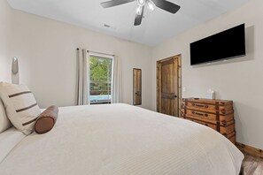 Une chambre à coucher avec un lit, un ventilateur de plafond, une télévision, une porte en bois et une fenêtre avec des rideaux.