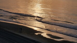 Ein Strand mit untergehendem Sonnenuntergang am Horizont, sanfte Wellen, die ans Ufer schlagen, und einige Personen, die am Strand entlanggehen.