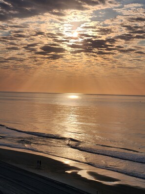 Uma praia com o sol se pondo no horizonte, ondas batendo suavemente na costa, e algumas pessoas caminhando pela areia.