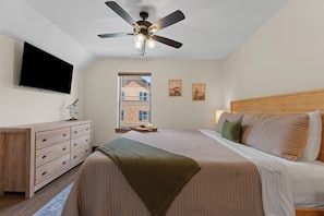 Ein Schlafzimmer mit einem Bett, einem Deckenventilator, einem Fernseher, einer Kommode und einem Fenster mit Blick auf ein Gebäude.
