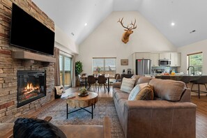 Amplia sala de estar con chimenea, cabeza de venado montada, televisor de pantalla plana y comedor con cocina al fondo.