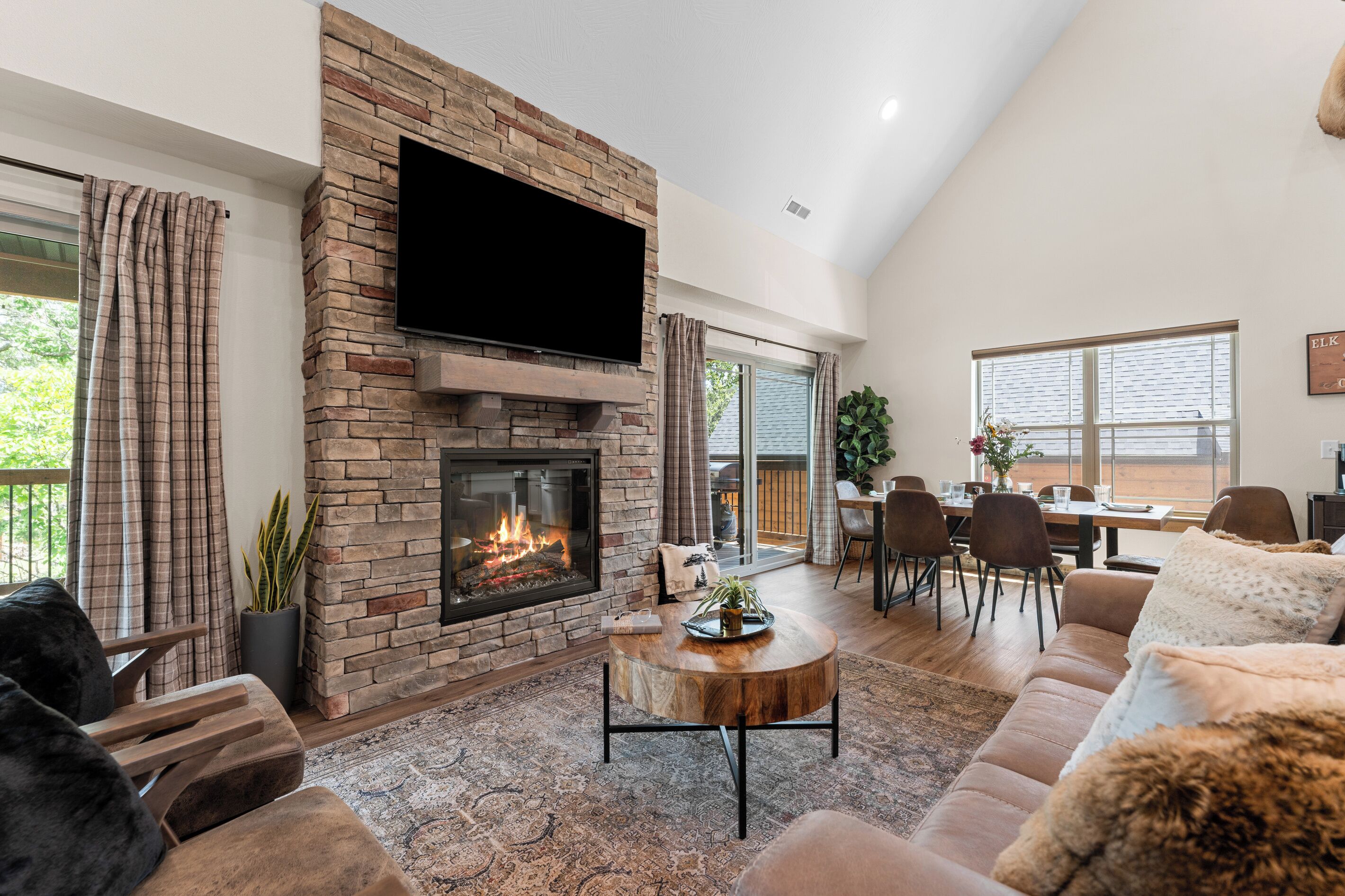 A spacious living room with a stone fireplace, a flat-screen TV mounted above it, a dining area with a table and chairs, and a large window providing a view of the outdoors.