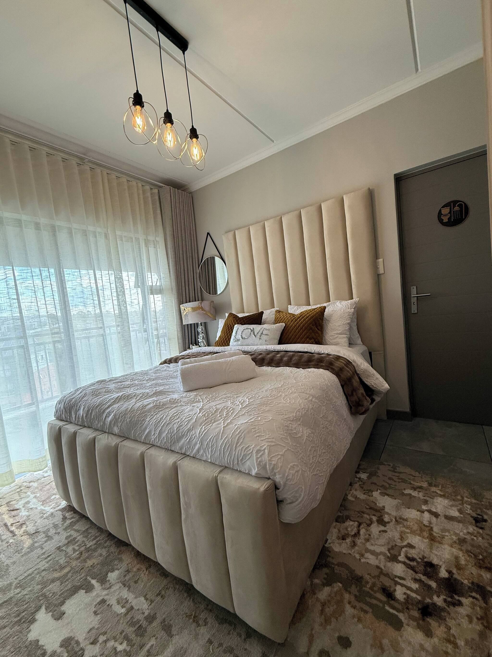 A modern bedroom with a large bed, a headboard, a bedside table, a mirror, and a door.