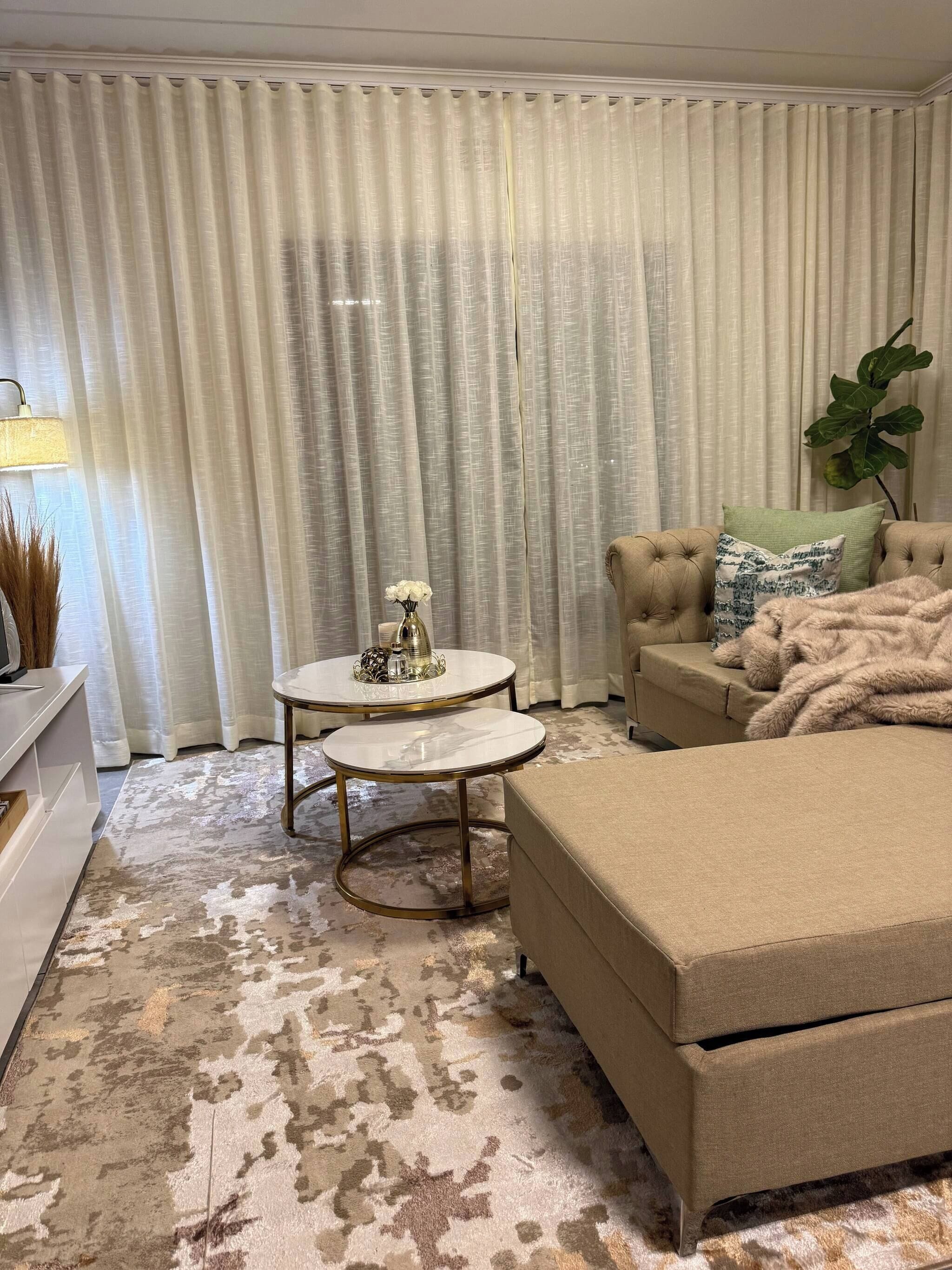 A living room with a beige sofa, a round coffee table, a white lamp, and a large window with sheer curtains.