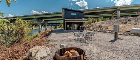 A modern outdoor seating area with a fire pit, a covered patio, and a view of a bridge.