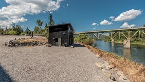 A gravel area with a small black building, a river, and a green bridge. - Everything, In Montana, All At Once (Saint Regis)