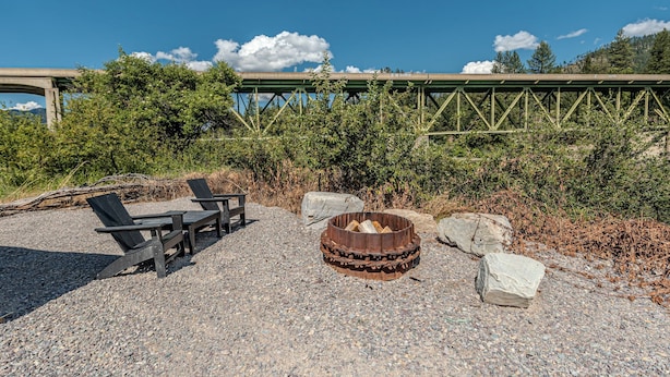 A gravel area with a fire pit and wooden chairs, a bridge in the background, and dense greenery. - Everything, In Montana, All At Once (Saint Regis)