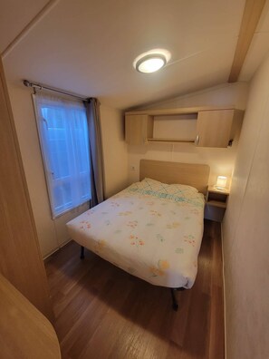 A small, cozy bedroom with a bed, a window with curtains, a ceiling light, and wooden furniture. - Air-conditioned mobile home at a 4-star campground for 6–8 people, with a pool, 5 minutes from the beaches (La Londe-les-Maures)