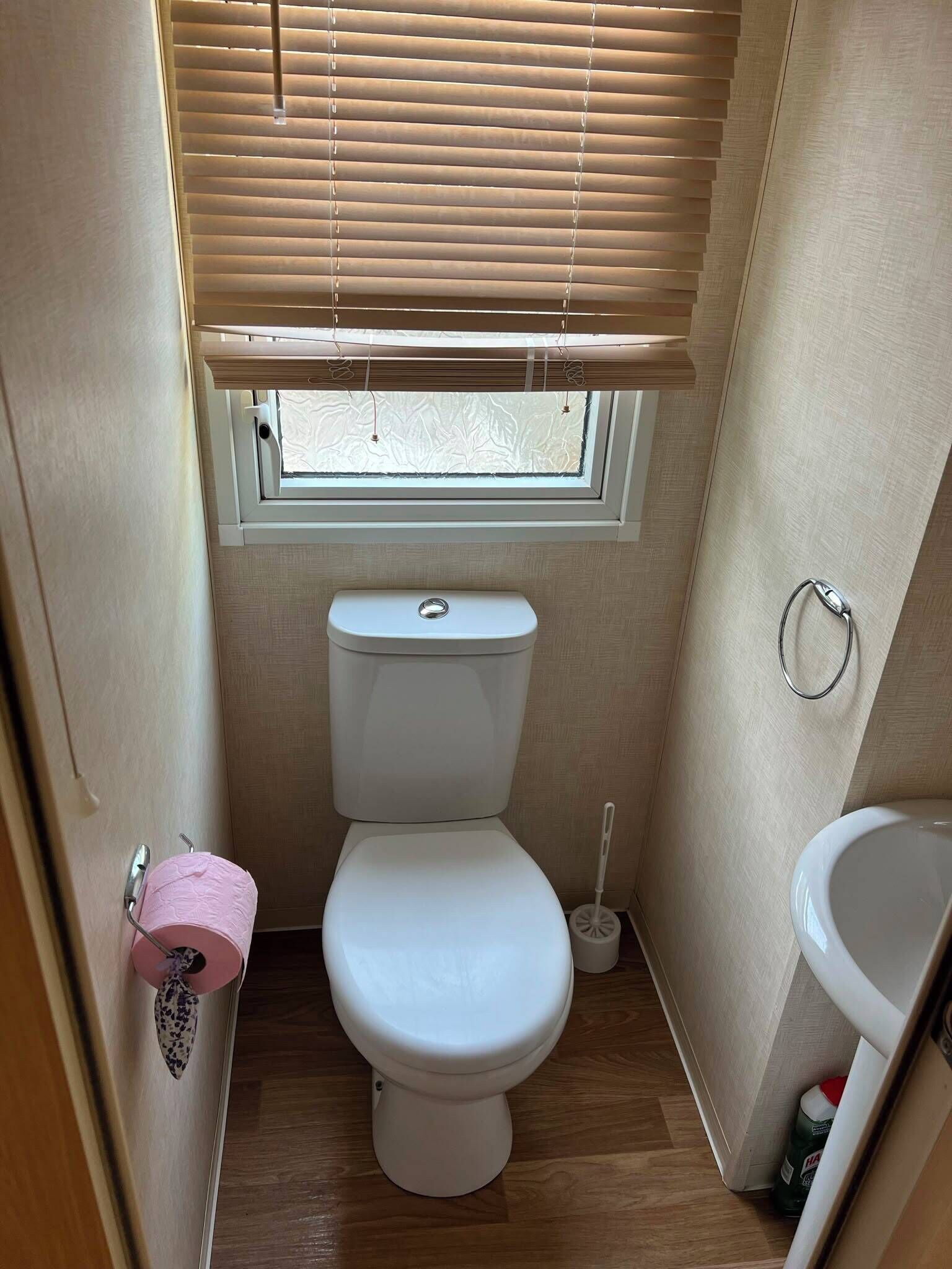 A compact bathroom with a toilet, a window with blinds, a toilet paper holder, and a sink.