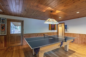 Ein Tischtennistisch in einem Raum mit Holzwänden und -decke, einer Tür, die nach draußen führt, und einem Dartboard an der Wand.