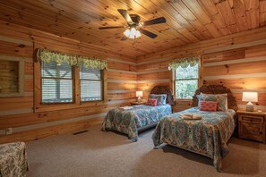Ein Schlafzimmer in einer Blockhütte mit zwei Betten, einem Deckenventilator und Fenstern mit Vorhängen.