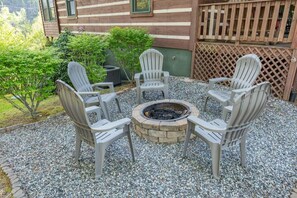 Ein kreisförmiger Sitzbereich mit vier Adirondack-Stühlen um ein Steinkaminfeuer, auf Kiesgrund mit Holzlattenzaun und Grünpflanzen im Hintergrund.