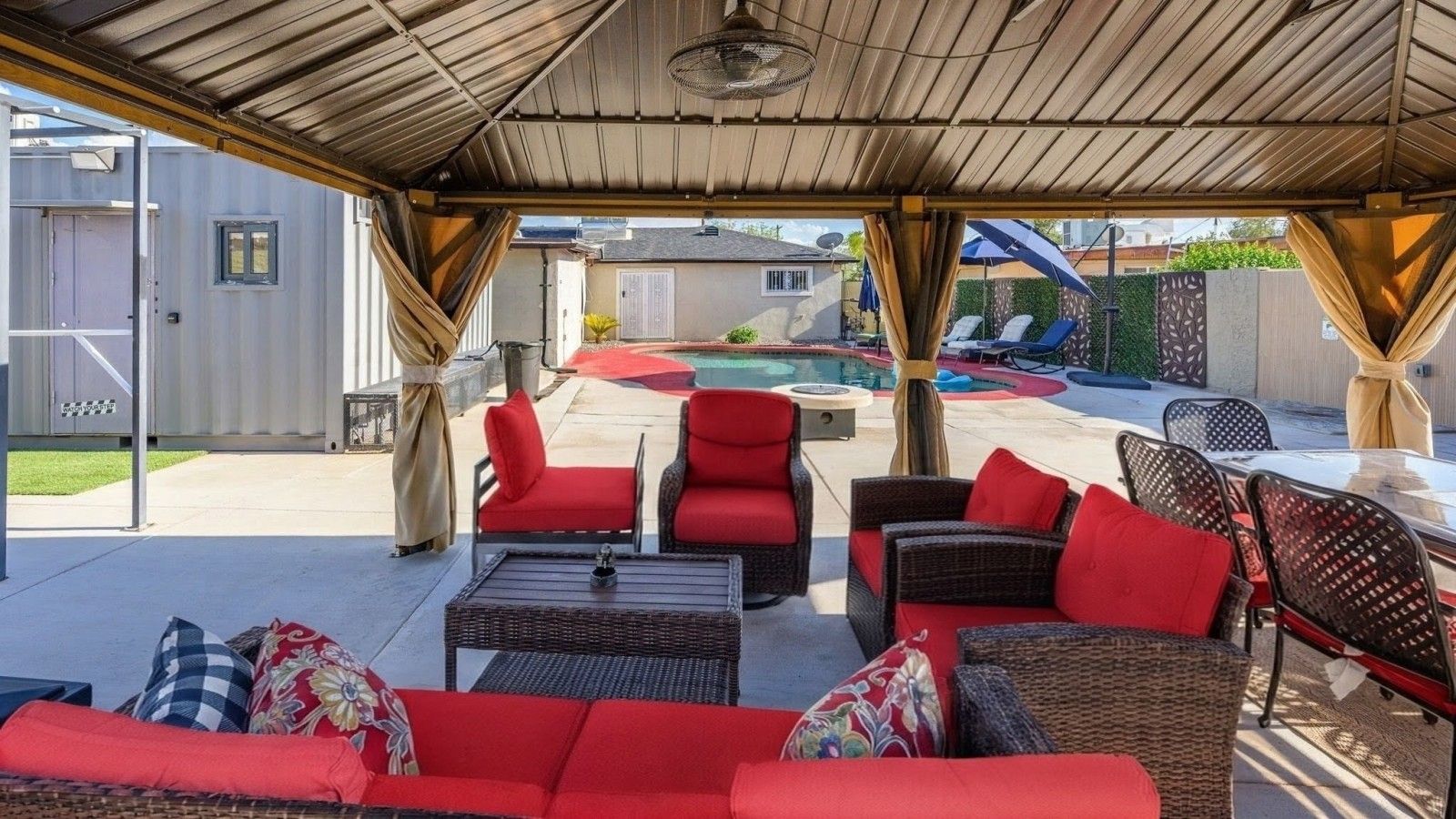 An outdoor patio with wicker furniture, red cushions, and a pool in the background.