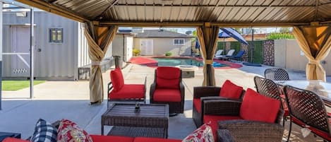 An outdoor patio with wicker furniture, red cushions, and a pool in the background.