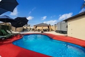 Ein Swimmingpool mit roter Liegefläche, Liegestühlen, Sonnenschirmen und einem überdachten Sitzbereich.