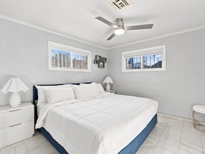 A bedroom with a bed, nightstands, a ceiling fan, and two windows with decorative ironwork.