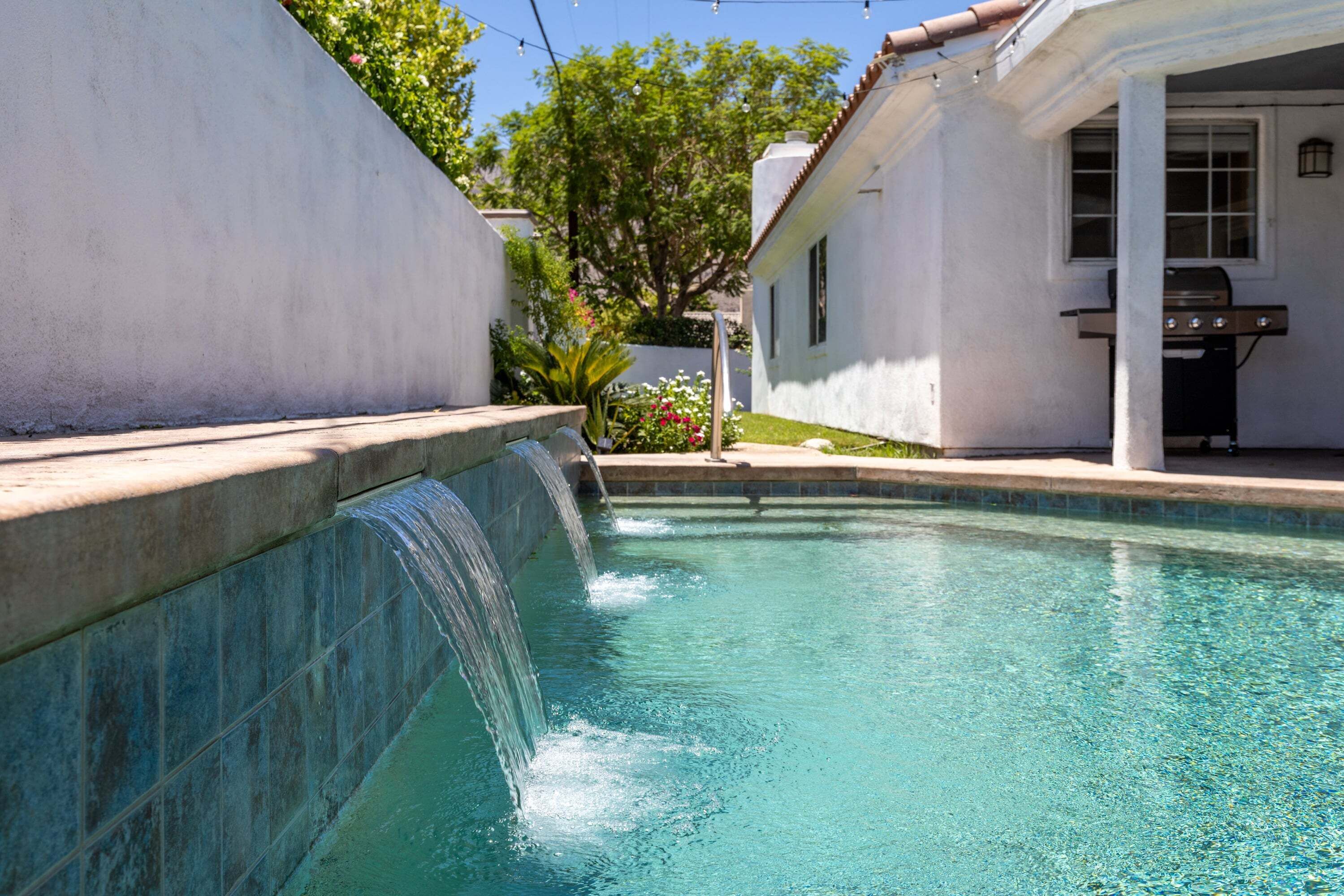 A swimming pool with a waterfall feature, a barbecue grill, and a white building with a window.