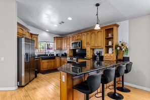 A modern kitchen with wooden cabinets, stainless steel appliances, a central island with bar stools, and recessed lighting. - The Cove Villa | 3BR | Private Pool | Sleeps 6 (La Quinta)
