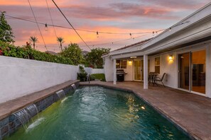 Ein Swimmingpool mit Wasserfall, eine Terrasse mit Gartenmöbeln und ein überdachter Bereich mit Grill.
