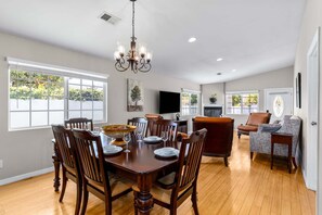 A spacious living room with a dining area, wooden floors, a chandelier, and large windows. - The Cove Villa | 3BR | Private Pool | Sleeps 6 (La Quinta)