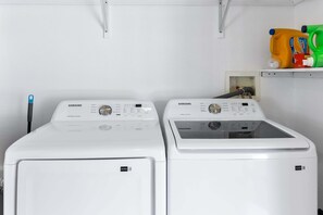Two white Samsung washing machines in a laundry room with a shelf above containing cleaning supplies. - The Cove Villa | 3BR | Private Pool | Sleeps 6 (La Quinta)