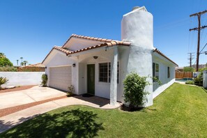 A single-story house with a white exterior, a garage, and a small front yard with a lawn and a few shrubs. - The Cove Villa | 3BR | Private Pool | Sleeps 6 (La Quinta)