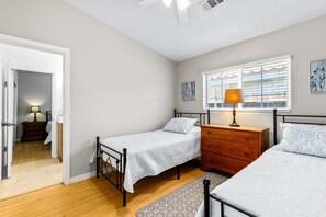 A bedroom with two single beds, a wooden dresser, a nightstand with a lamp, and a window with blinds. - The Cove Villa | 3BR | Private Pool | Sleeps 6 (La Quinta)