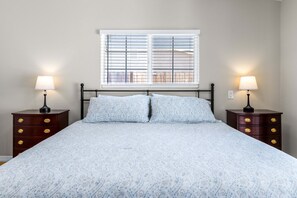 Une chambre à coucher avec un lit, deux tables de chevet, une fenêtre avec des stores et une vue sur une clôture.