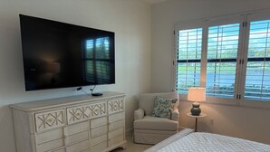 A bedroom with a bed, a TV mounted on a dresser, a chair, a window with blinds, and a lamp on a side table. - Renovated Ravenna first floor townhome in exclusive Pelican Marsh. (Naples)