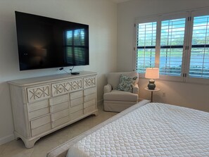 A bedroom with a bed, a TV mounted on a dresser, a chair, a window with blinds, and a lamp on a side table. - Renovated Ravenna first floor townhome in exclusive Pelican Marsh. (Naples)