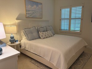 A bedroom with a bed, a nightstand, a lamp, a digital clock, a painting, and a window with blinds. - Renovated Ravenna first floor townhome in exclusive Pelican Marsh. (Naples)