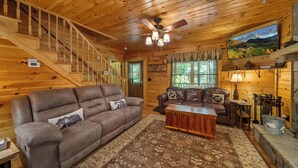 A wooden cabin interior with a sofa, a coffee table, a fireplace, a television, and a staircase. - Classic Log Cabin with Hot Tub & Fire Pit (West Jefferson)