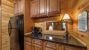 A kitchen with wooden cabinets, a black refrigerator, a coffee maker, and a sign with a message. - Classic Log Cabin with Hot Tub & Fire Pit (West Jefferson)