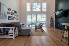 A spacious living room with a large window, a gray sectional sofa, a wooden coffee table, a wooden chair, and a flat-screen TV mounted on a dark wall. - Beautiful Bungalow - Steps from Okanagan Lake! Luxurious and family-friendly. (Kelowna)
